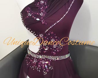 Puede incluir: Un traje de baile de color morado oscuro con escote asimétrico, doble tirante y una abertura lateral. La prenda está adornada con intrincados apliques de encaje, cristales transparentes dispersos y una falda de tela semitransparente.