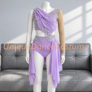 May include: A lavender dance costume featuring a one-shoulder design with a sheer nude sleeve, embellished with silver rhinestones and floral appliqués. The bodice includes draped fabric, and the skirt consists of layered chiffon panels.