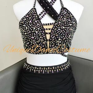 May include: Black dance costume with sparkling embellishments. The halter top and skirt are adorned with iridescent rhinestones. The top features a crisscross strap design and cut-out detail. The skirt has a draped overlay.