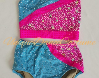 May include: A two-piece dance costume in turquoise and hot pink. The top is a one-shoulder design, and the bottoms are high-waisted. Both pieces are embellished with sparkling rhinestones. The text "Uniques Dance Costume" is visible.
