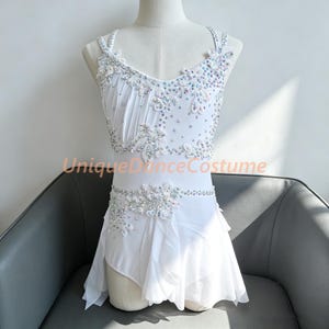 May include: A white dance costume featuring a sleeveless bodice with thin straps, adorned with intricate white floral lace appliques and scattered iridescent crystals. The skirt consists of layered sheer white fabric with a ruffled hemline.