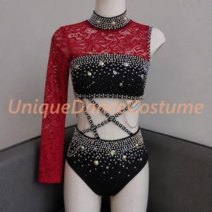 May include: A black and red dance costume featuring a high-neck lace bodice with one long sleeve, a cropped top, and high-waisted bottoms. The garment is embellished with numerous silver studs, rhinestones, and thin chains. Text reads UniqueDanceCostume.