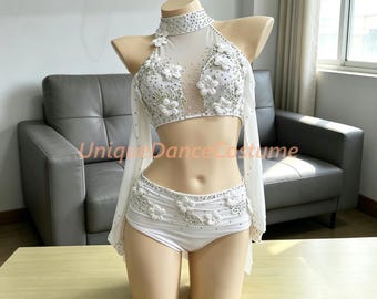 May include: A white two-piece dance costume featuring a high-neck crop top and matching bottoms. The set is adorned with white fabric flowers, clear rhinestones, and sheer mesh panels. Long, flowing sheer white sleeves are attached to the top.
