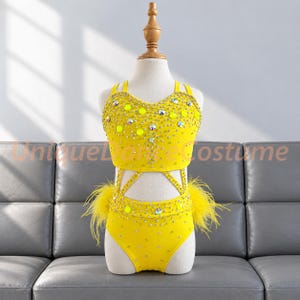 May include: A bright yellow two-piece dance costume featuring a crop top and high-waisted bottoms. The set is heavily embellished with clear crystals, iridescent sequins, and yellow feathers on the sides. The text UniqueDanceCostume is visible.