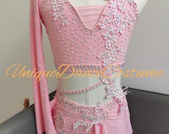 May include: Pink dance costume with rhinestones and floral lace appliques. The costume features a halter top with one long sleeve, a matching shorts set, and a flowing skirt overlay. The text "Unique Dance Costume" is visible on the costume.