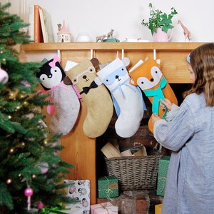 May include: Five felt Christmas stockings hanging on a fireplace mantel. The stockings are decorated with animal faces and have a variety of colors, including gray, brown, blue, and orange.