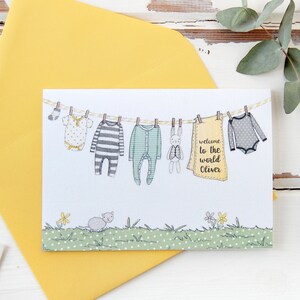 A personalised new baby greetings card with a watercolour painted illustration of a washing line full of baby clothes, lays on a yellow envelope. Both are on a white desk.