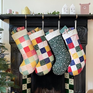 Puede incluir: Cuatro calcetines de patchwork coloridos cuelgan de una repisa de chimenea negra. Cada calcetín está lleno de regalos envueltos. Los calcetines están decorados con una variedad de colores, incluyendo rojo, verde, azul, amarillo y rosa.