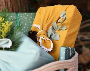 Op de afbeelding: Een bruine geweven mand gevuld met geschenken verpakt in gele stof met witte kant en groene bladeren, en een groen-witte gepatroneerde geschenkdoos.