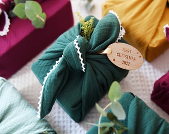 Op de afbeelding: Een groen stoffen cadeau verpakt met een witte kantversiering en een houten label met de tekst "First Christmas 2022".