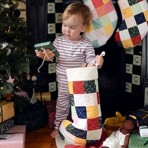 May include: A child holds a wrapped gift in front of a Christmas tree and a patchwork stocking. The stocking is red, green, blue, and pink with white polka dots. The child is wearing a striped sweater.