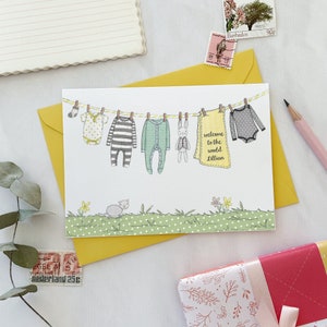 A personalised new baby greetings card with a watercolour painted illustration of a washing line full of baby clothes, lays on a yellow envelope. Both are on a white desk.