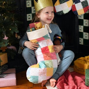 May include: A child wearing a blue and white pyjama top with a crown on their head holds a patchwork Christmas stocking filled with gifts. The stocking is made of colourful squares with a white quilted design.