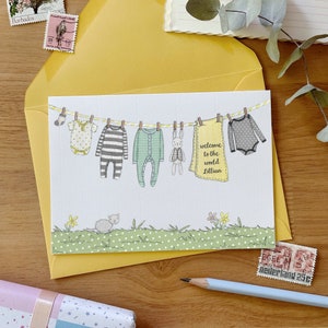 A personalised new baby greetings card with a watercolour painted illustration of a washing line full of baby clothes, lays on a yellow envelope. Both are on a wooden desk.