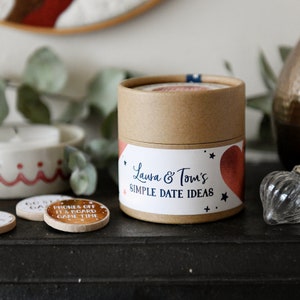 Circular Cardboard carton with a paper sticker on the front is placed on the mantle of a black fireplace. Next to the carton is 2 wooden disks with copper foiled stickers attached to the top of them.