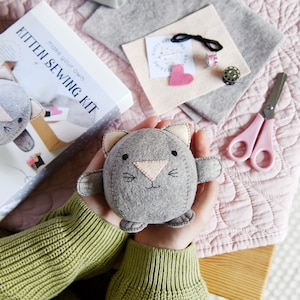 Kitten sewing kit is placed on a pink blanket next to different coloured felt pieces, two metal cotton spools and a pair of pink scissors. There is a person holding a round felt grey kitten in their hands. The person is wearing a green jumper.
