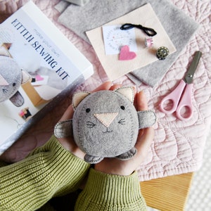 Kitten sewing kit is placed on a pink blanket next to different coloured felt pieces, two metal cotton spools and a pair of pink scissors. There is a person holding a round felt grey kitten in their hands. The person is wearing a green jumper.