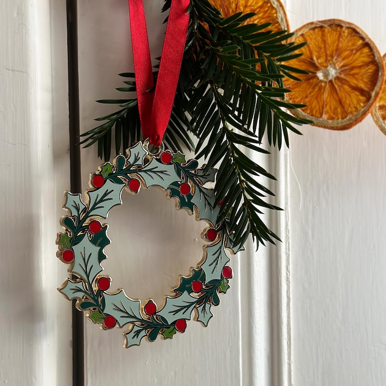 An heirloom Christmas wreath decoration with holly leaves and red berries hangs from a red ribbon on a white door handle with festive greenery and orange slice garland.