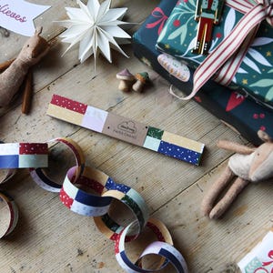 A set of traditional patchwork paper chains lays on a wooden table beside a stack of wrapped Christmas gifts, two small reindeer toys, and a paper star decoration. A made up chain of paper chains lays in front of the pack.
