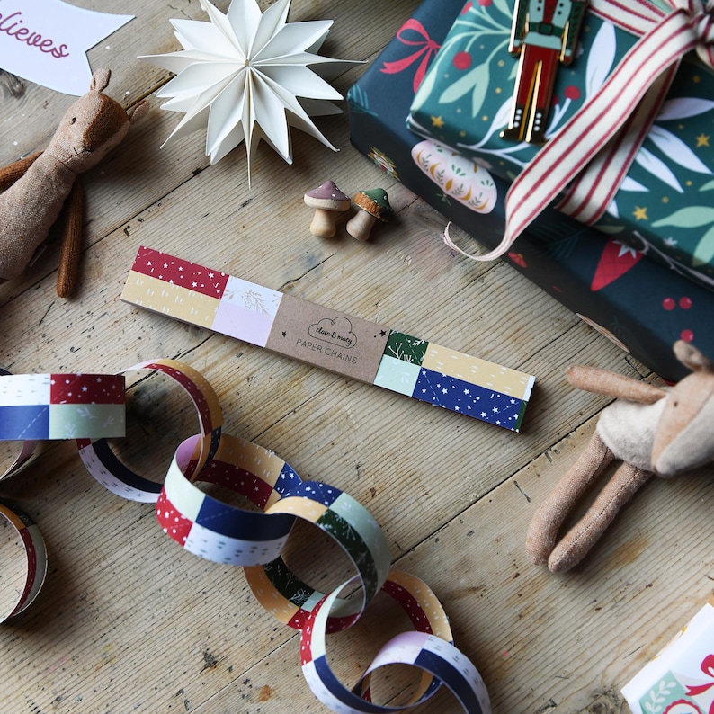 A set of traditional patchwork paper chains lays on a wooden table beside a stack of wrapped Christmas gifts, two small reindeer toys, and a paper star decoration. A made up chain of paper chains lays in front of the pack.