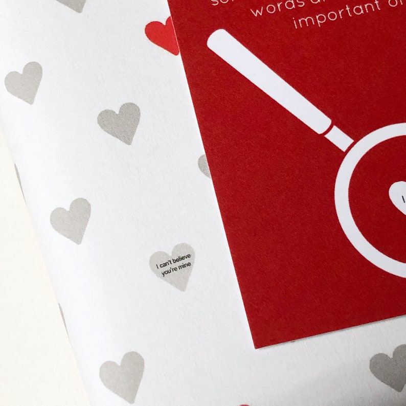 Tiny Heart Valentines Gift Wrap. White gift wrap featuring tiny grey and red hearts, some with tiny messages in is used to wrap a gift and finished off with a red bow.