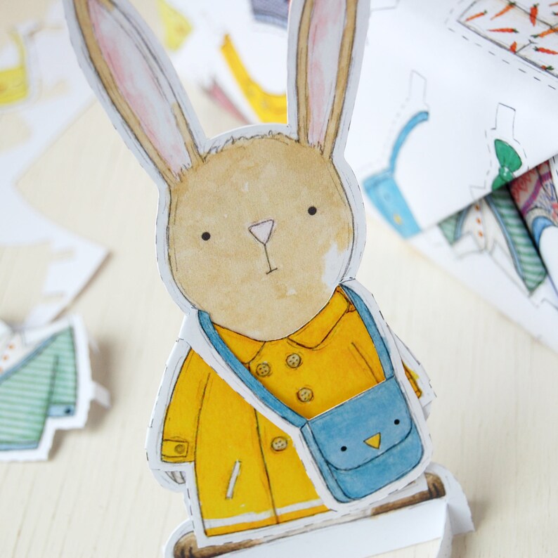 Puede incluir: Una mu&ntilde;eca de papel de un conejo marr&oacute;n que lleva un impermeable amarillo y una bolsa azul con un dise&ntilde;o de p&aacute;jaro.