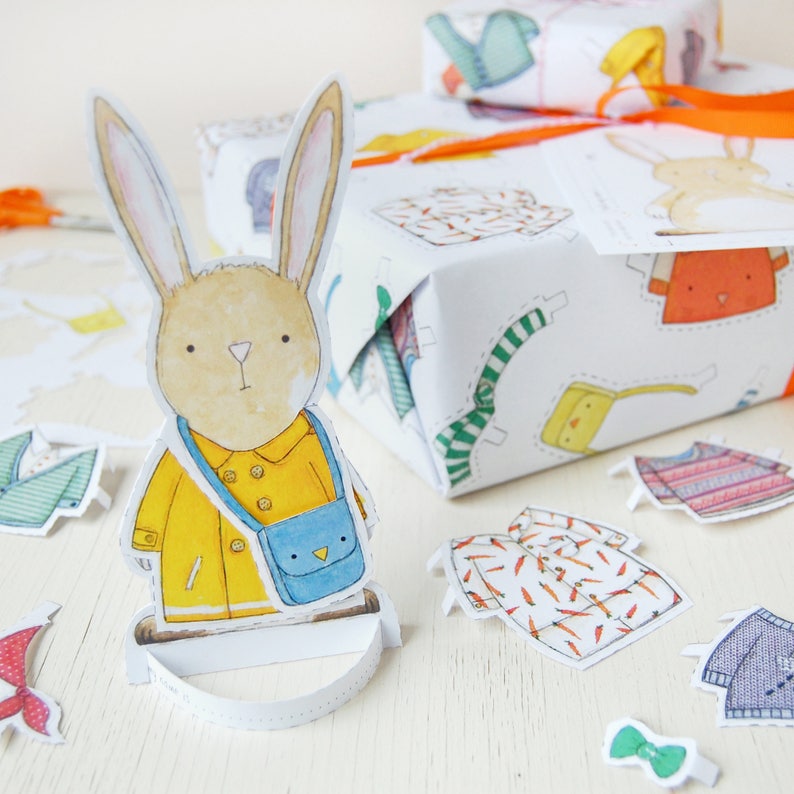 Puede incluir: Un conejo de papel artesanal que lleva un impermeable amarillo con una bolsa azul. El conejo est&aacute; de pie sobre una base blanca. El fondo incluye una caja de regalo blanca con un patr&oacute;n colorido de ropa.