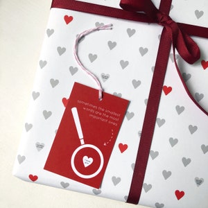 Tiny Heart Valentines Gift Wrap. White gift wrap featuring tiny grey and red hearts, some with tiny messages in is used to wrap a gift and finished off with a red bow.