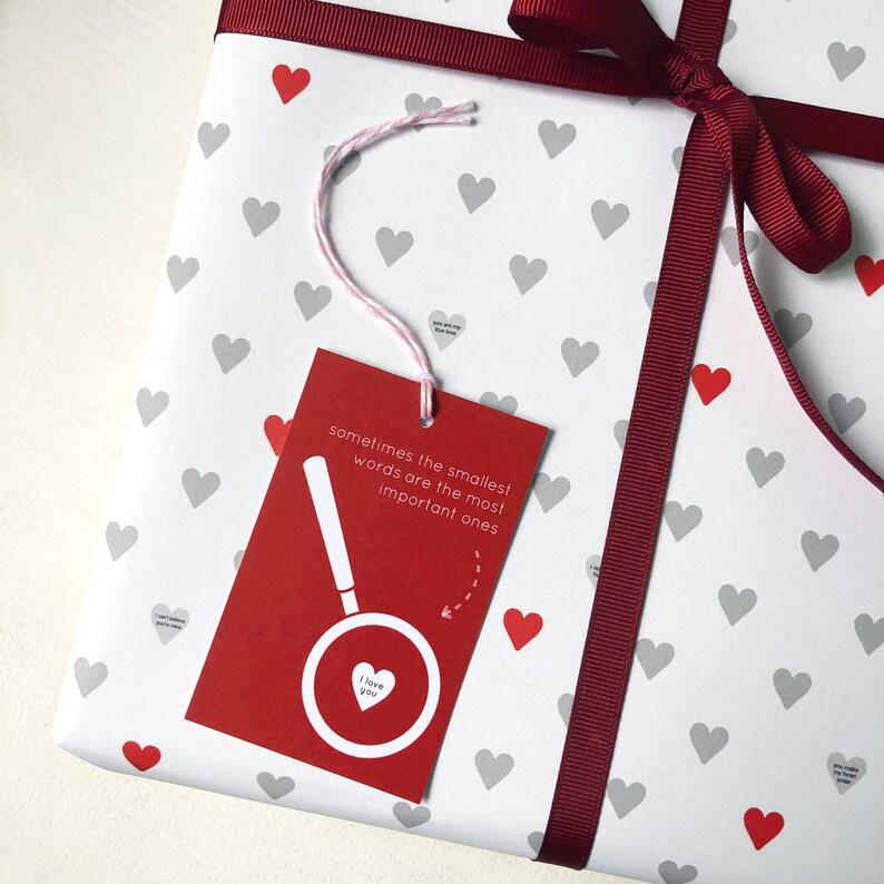 Tiny Heart Valentines Gift Wrap. White gift wrap featuring tiny grey and red hearts, some with tiny messages in is used to wrap a gift and finished off with a red bow.