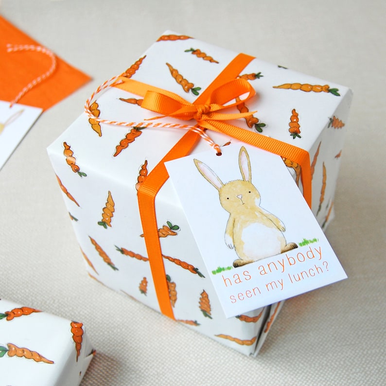 May include: A white gift box with an orange ribbon and a gift tag. The gift box is wrapped in paper with a pattern of orange carrots. The gift tag has a cartoon illustration of a brown rabbit with a white belly and the text "has anybody seen my lunch?"