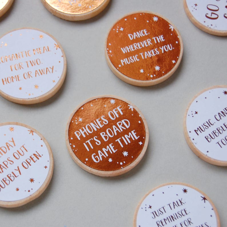 15 wooden disks scattered on a grey background. 7 of the wooden disks have white stickers with copper foiled writing on them, 8 of the disks have copper stickers with white writing on them