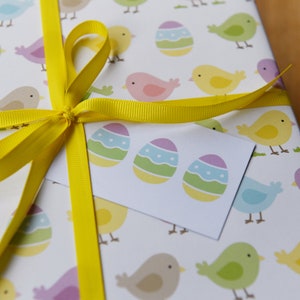 May include: A white gift bag with a yellow ribbon bow. The bag is decorated with a repeating pattern of colorful Easter eggs and birds. A white gift tag with three colorful Easter eggs is attached to the bag.