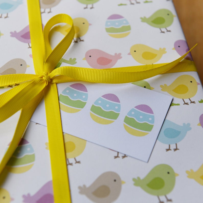 May include: A white gift bag with a yellow ribbon bow. The bag is decorated with a repeating pattern of colorful Easter eggs and birds. A white gift tag with three colorful Easter eggs is attached to the bag.