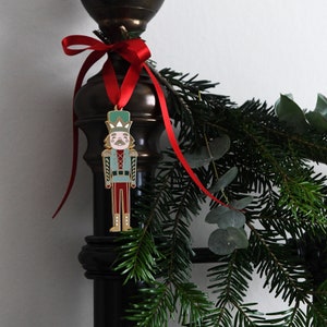 A gold, red, and green enamel Nutcracker Christmas tree decoration hangs from a red ribbon on a door handle, on a black bed post.