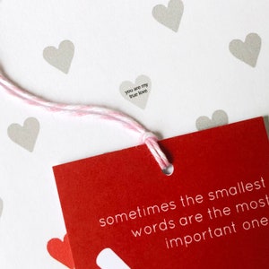 Tiny Heart Valentines Gift Wrap. White gift wrap featuring tiny grey and red hearts, some with tiny messages in is used to wrap a gift and finished off with a red bow.