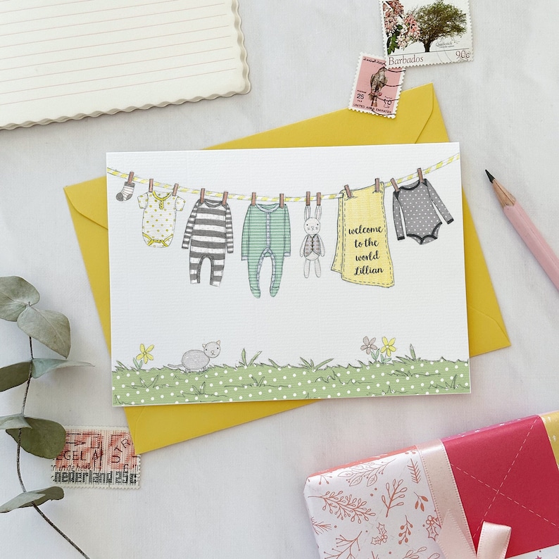 A personalised new baby greetings card with a watercolour painted illustration of a washing line full of baby clothes, lays on a yellow envelope. Both are on a white desk.