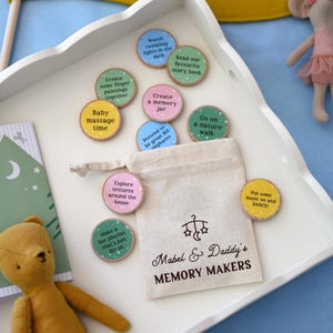Può includere: Un vassoio bianco contiene dischi di legno colorati con suggerimenti di attività come "Baby massage time" e "Go on a nature walk". Una piccola borsa di tela recita "Mabel & Daddy's MEMORY MAKERS".