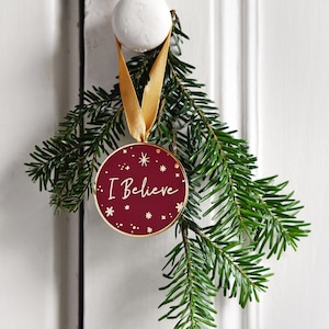 May include: A gold-colored Christmas ornament with a red background and the words "I Believe" in white script. The ornament is hanging from a gold ribbon and is surrounded by green pine branches.