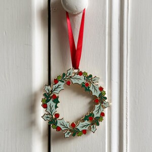An heirloom Christmas wreath decoration with holly leaves and red berries hangs from a red ribbon on a white door handle.