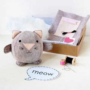 Hand sewn grey felt kitten is placed in front of a craft kit box filled with felt, cotton and stuffing.