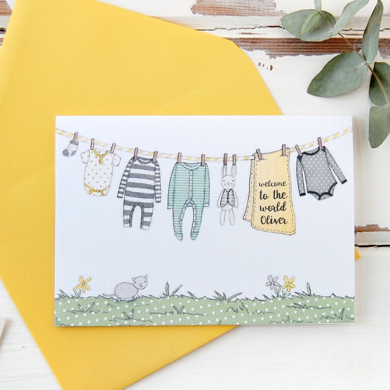 A personalised new baby greetings card with a watercolour painted illustration of a washing line full of baby clothes, lays on a yellow envelope. Both are on a white desk.