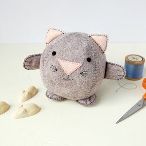 A Hand sewn grey felt kitten is placed next to a spool of cotton a pair of scissors and some white chocolate mice