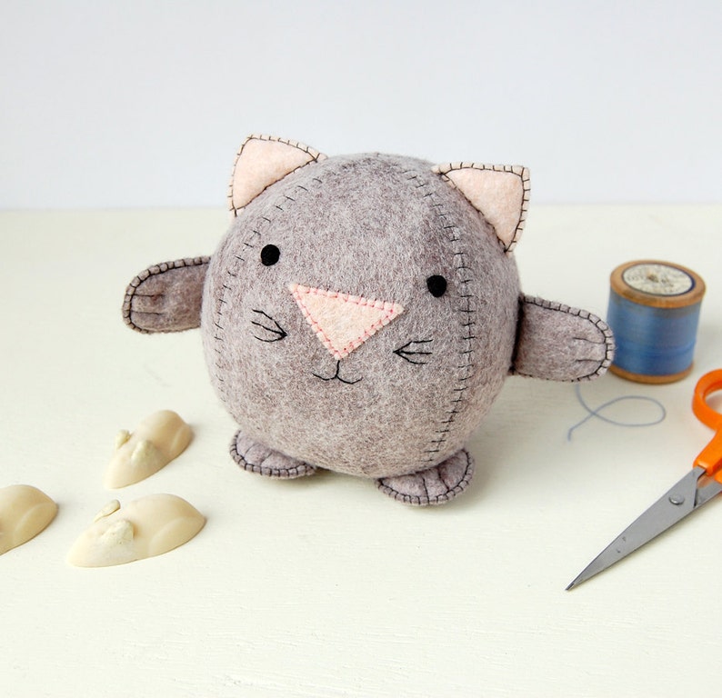 A Hand sewn grey felt kitten is placed next to a spool of cotton a pair of scissors and some white chocolate mice