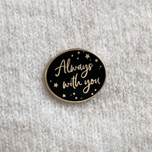 Black oval pin badge with gold ring and small gold stars reading always with you in a gold handwriting font on a cream knitted jumper.