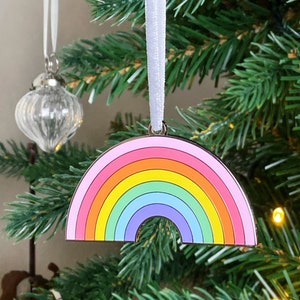 May include: A rainbow-colored Christmas ornament shaped like a rainbow. The ornament is made of metal and has a gold finish. It is hanging from a white ribbon on a Christmas tree.