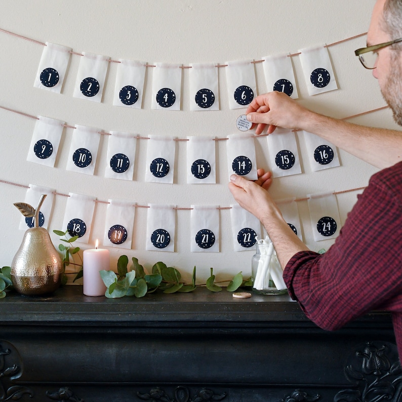 Three rows of white envelopes, each with a numbered sticker on, strung on ribbon are hung on a neutral wall above a black mantlepiece. Two hands are reaching in to open one of the envelopes.