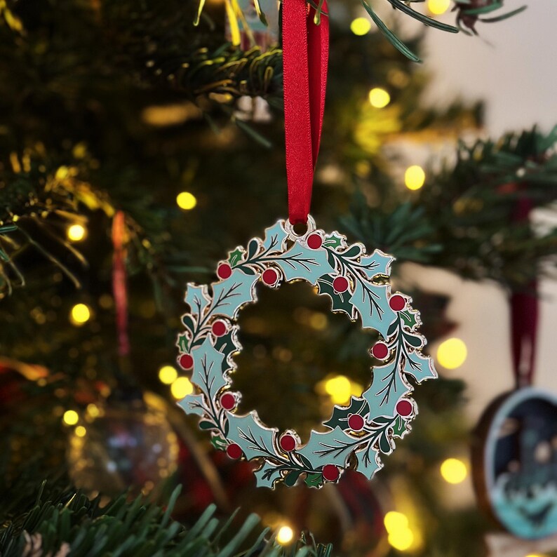 An heirloom holly wreath Christmas decoration hangs from a red ribbon on a decorated Christmas tree.