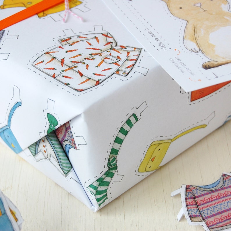 Puede incluir: Papel de regalo blanco con un dise&ntilde;o recortado de varias prendas de vestir, incluyendo una bufanda a rayas, un pollito amarillo, una camisa con estampado de zanahorias y un conejito. El texto "&iexcl;Estoy listo para la fiesta!" est&aacute; impreso en el papel.
