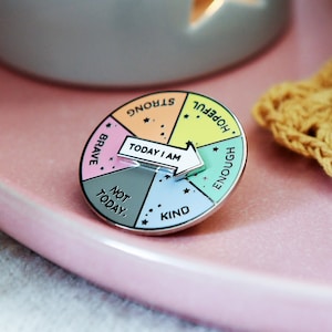 Rainbow pin badge with the words strong, hopeful, enough, kind, not today and brave on with a white and silver spinner in the centre reading 'today i am' so you can choose your mood. Pin on pink plate near candle and crocheted yellow coaster