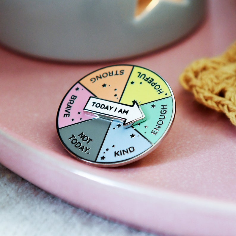 Rainbow pin badge with the words strong, hopeful, enough, kind, not today and brave on with a white and silver spinner in the centre reading 'today i am' so you can choose your mood. Pin on pink plate near candle and crocheted yellow coaster